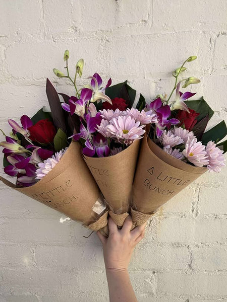 Bouquet of flowers wrapped in brown paper with 'A Little Bunch' branding against a white brick wall.