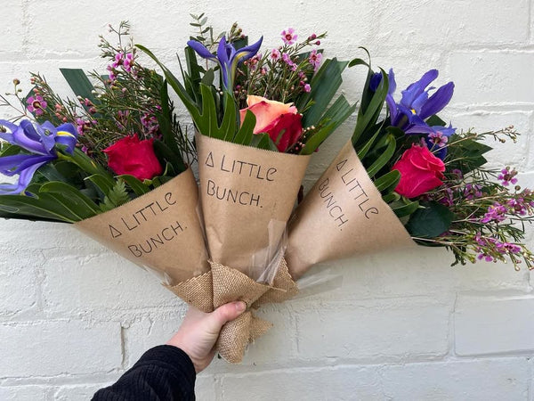 Bouquet of flowers held by a hand with 'A Little Bunch' packaging against a white brick wall.